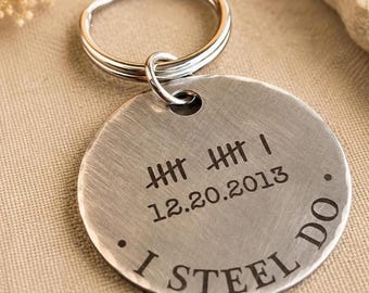 May include: A round, brushed metal keychain featuring an engraved tally mark design, the date 12.20.2013, and the text I STEEL DO. The circular metal tag is attached to a standard silver-colored metal split ring for keys.