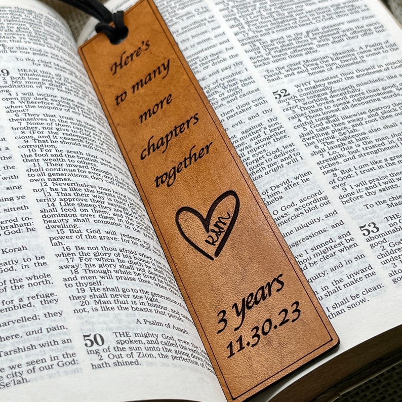 May include: A brown leather bookmark with the text "Here's to many more chapters together" and a heart with the initials "R&M" engraved. The bookmark also includes the text "3 years 11.30.23".