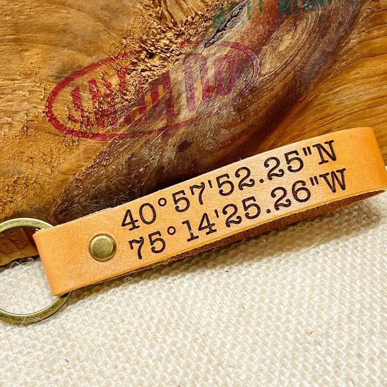 May include: A tan leather keychain with a brass ring and a metal rivet. The keychain has the coordinates "40&deg; 57' 52.25" N 75&deg; 14' 25.26" W" engraved on it.