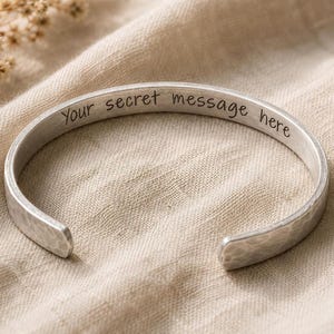 May include: A silver-colored cuff bracelet with the engraved text "Your secret message here." The bracelet has a hammered texture and an open design. The bracelet is displayed on a beige fabric surface.