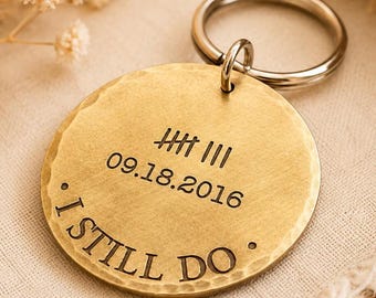 May include: A round, gold-colored keychain with the text "I STILL DO" and the date 09.18.2016 engraved on it. The keychain has a silver-colored ring attached. The surface of the keychain has a hammered texture.
