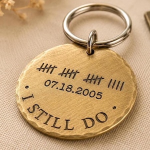 May include: A round, gold-colored metal keychain tag with the words "I STILL DO" engraved at the bottom. The tag also features the date "07.18.2005" and tally marks. A silver-colored key ring is attached at the top.