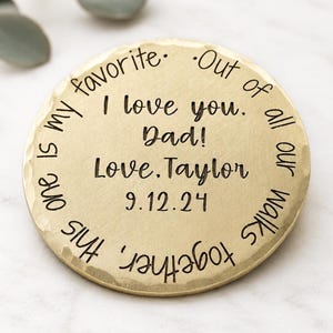 May include: A round, gold-colored metal token with a hammered edge. The center is engraved with the text: I love you. Dad! Love, Taylor 9.12.24. The outer rim features the engraved text: Out of all our walks together, this one is my favorite.