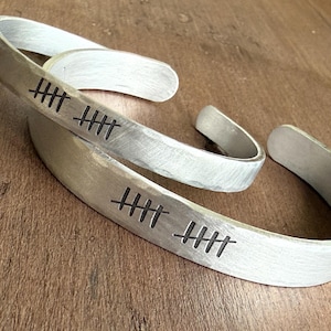 May include: Three silver-toned metal bracelets with a brushed finish. Each bracelet has a series of etched tally marks. The bracelets are displayed on a wooden surface. The bracelets are open cuff style.