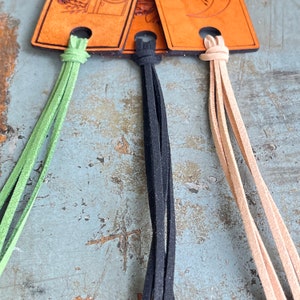 May include: Three leather bookmarks with different colored tassels. The bookmarks are brown with a design on the front. The tassels are green, black, and beige.