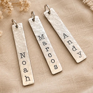 May include: Three rectangular silver-toned metal pendants with a hammered texture, each featuring a jump ring at the top. The pendants are stamped with the names Noah, Marcos, and Andy in black capital and lowercase letters arranged vertically.