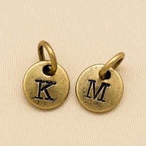 May include: Two circular metal charms with a distressed antique brass finish. Each charm features a single stamped capital letter, one displaying the letter K and the other displaying the letter M, with an attached jump ring at the top of each piece.