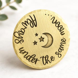 May include: A circular gold-toned metal disc with a hammered edge. The surface is engraved with the text "Always under the same moon..." in a cursive font, surrounding a central graphic of a crescent moon and two small stars.