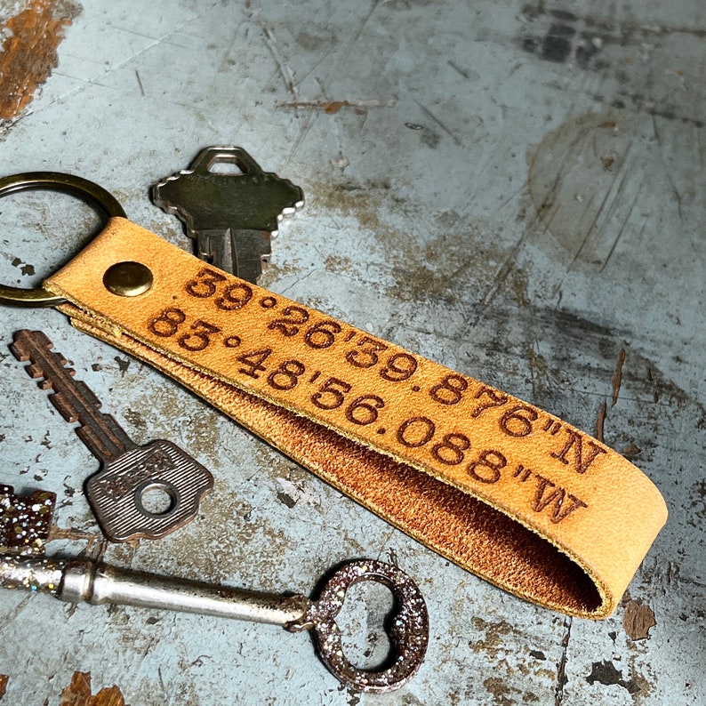 May include: A tan leather keychain with the coordinates "39.2639.876"N 85.4856.088"W" engraved on it. The keychain is attached to a silver key.