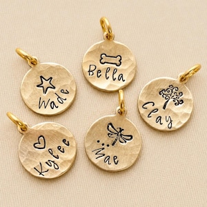 May include: Five round gold-toned metal charms with a hammered texture, each featuring a jump ring at the top. The charms are stamped with black ink designs and names: a star with Wade, a bone with Bella, a tree with Clay, a heart with Kylee, and a dragonfly with Mae.