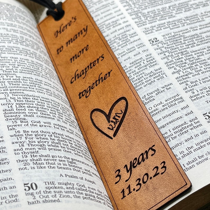 May include: A brown leather bookmark with the text "Here's to many more chapters together" and a heart with the initials "R&M" inside. The bookmark also has the text "3 years 11.30.23" engraved on it.