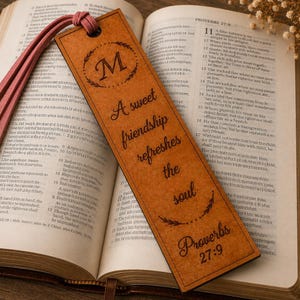 May include: A rectangular brown wooden bookmark featuring a laser-engraved letter M inside a laurel wreath, the text A sweet friendship refreshes the soul, and the citation Proverbs 27:9, finished with a pink leather tassel at the top.