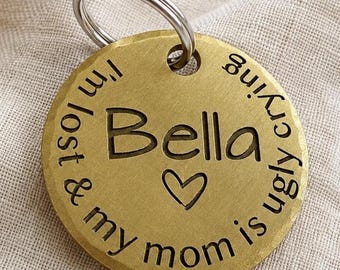 May include: A circular brass pet ID tag with a silver-toned metal split ring. The tag features the engraved name Bella, a heart symbol, and the text I'm lost & my mom is ugly crying, all centered on the front surface of the metallic disc.