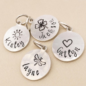 May include: Four circular silver-toned metal charms with a hammered texture. Each charm features a stamped name and a unique symbol: Kinley with a sun, Jackson with a four-leaf clover, Layne with a dragonfly, and Gatlyn with a heart symbol.