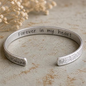 May include: Silver-toned cuff bracelet with the inscription "Forever in my heart." The open-ended bracelet features a floral design on the edges. The bracelet is displayed on a neutral-toned surface.