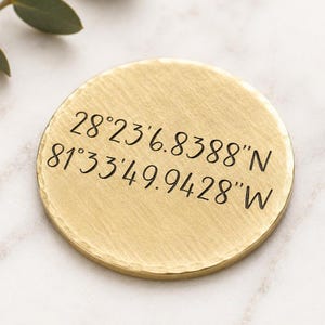 May include: A circular gold-toned metal disc with a hammered edge and a brushed finish. The surface is engraved with the black text 28°23'6.8388"N 81°33'49.9428"W, representing specific geographic coordinates in a clear, sans-serif font style.