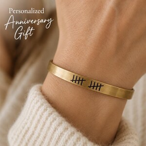 May include: A brushed gold-tone metal cuff bracelet featuring a black engraved tally mark design. The text "Personalized Anniversary Gift" is visible in the upper left corner of the frame. The bracelet has a smooth, flat, and open-ended band.