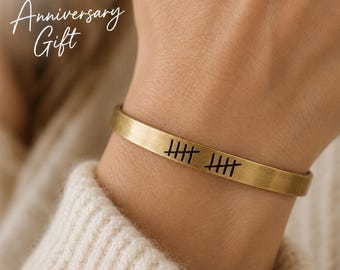 May include: A brushed gold-tone metal cuff bracelet featuring a black engraved tally mark design. The text "Personalized Anniversary Gift" is visible in the upper left corner of the frame. The bracelet has a smooth, flat, and open-ended band.