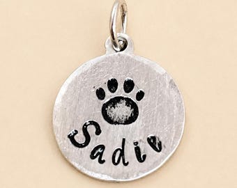 May include: A circular silver-toned metal charm featuring a stamped black paw print design above the name Sadie in a cursive font. The charm includes a small silver jump ring attached to the top for hanging, with a brushed metallic finish.