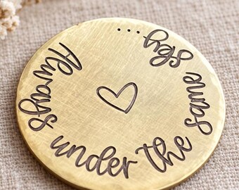 May include: A circular gold-toned metal disc with a hammered texture. The surface features the engraved text Always under the same sky... arranged in a circular pattern around a central heart symbol, creating a decorative and sentimental piece.