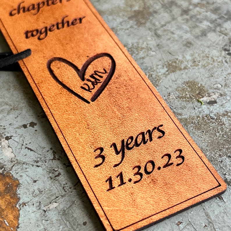 May include: A brown leather bookmark with the text "chapter together" and a heart with the word "rum" inside. The bookmark also has the text "3 years 11.30.23" engraved on it.