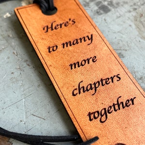 May include: A brown leather bookmark with the text "Here's to many more chapters together" engraved on it.