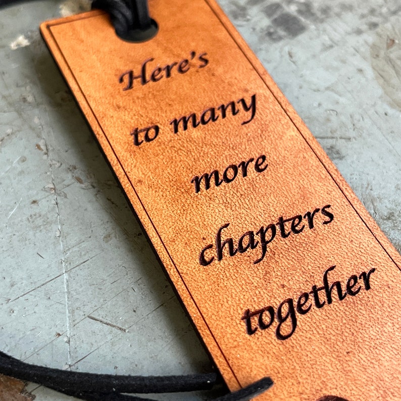 May include: A brown leather bookmark with the text "Here's to many more chapters together" engraved on it.