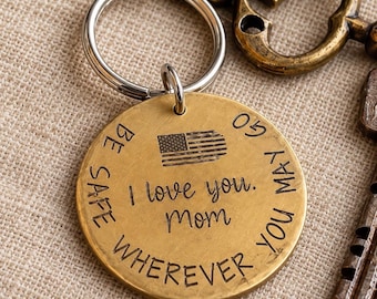 May include: A round brass keychain tag featuring an engraved American flag and the text "Be safe wherever you may go" and "I love you. Mom" in black lettering. The tag is attached to a silver-colored metal split ring.