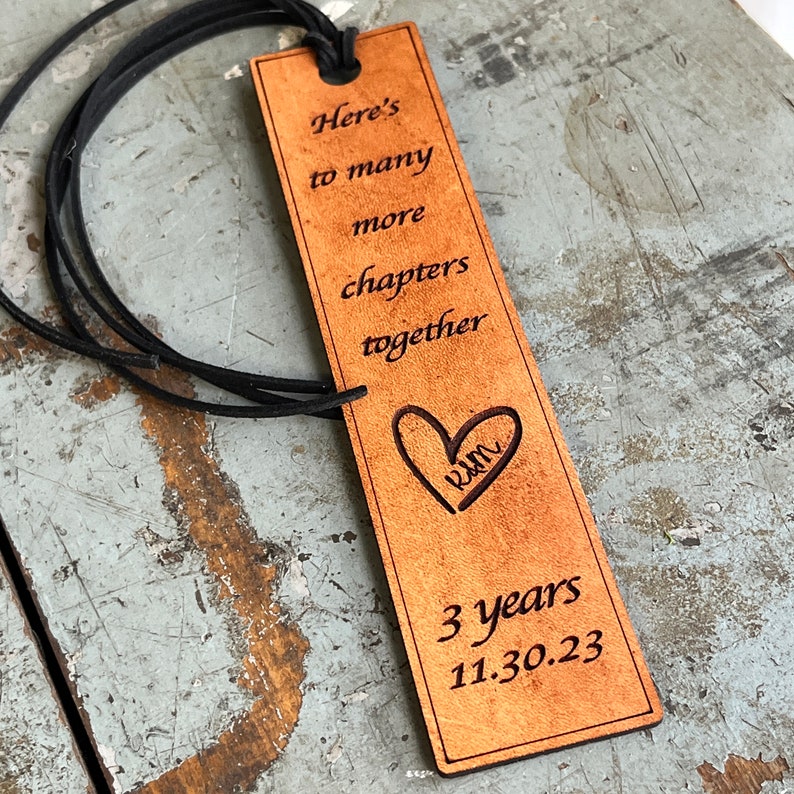 May include: A brown leather bookmark with the text "Here's to many more chapters together" and a heart with the initials "K&M" inside. The bookmark also has the text "3 years 11.30.23" on it.