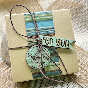 May include: A small, light brown gift box with a blue and green striped paper design. The box is tied with brown twine and has a wooden tag that says "For You" and a round wooden tag that says "Kylee Mae Designs".