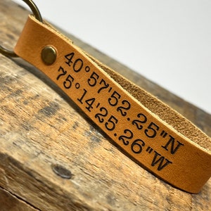May include: A brown leather keychain with a metal ring and a brass rivet. The keychain is engraved with the coordinates "40.57'52.25"N 75.14'25.26"W".