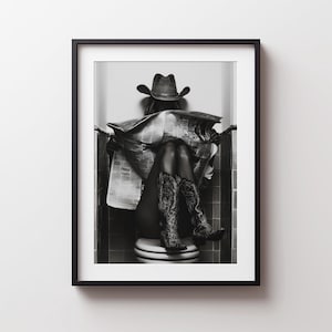 Può includere: Una stampa artistica incorniciata in bianco e nero che raffigura una figura con cappello da cowboy e stivali western a motivi, seduta su un water mentre legge un giornale. La composizione è centrata in una cornice nera con un passe-partout.