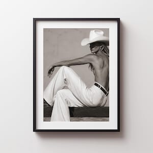 Può includere: Una stampa artistica incorniciata in bianco e nero che ritrae una persona con un cappello da cowboy a tesa larga e pantaloni chiari. Il soggetto è seduto di spalle, con la schiena scoperta e una cintura scura intorno alla vita.