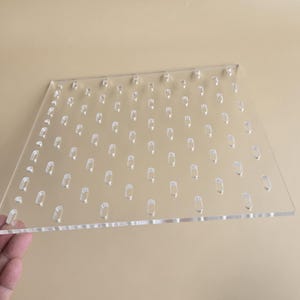 May include: Clear acrylic display panel with numerous small, evenly spaced, rectangular cutouts. The panel is held by a hand, showcasing its transparency and design for displaying small items. The background is a neutral beige.