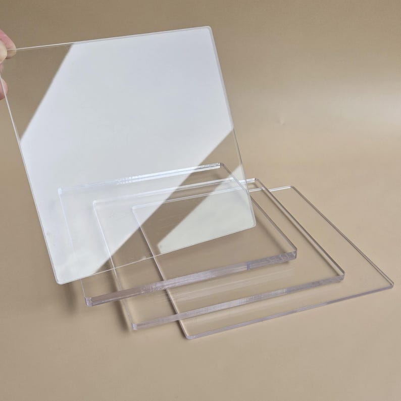 May include: A stack of clear acrylic sheets of varying sizes. The largest sheet is held by a hand, with a square shape and a slightly frosted appearance. The other sheets are rectangular and transparent, with clean edges.