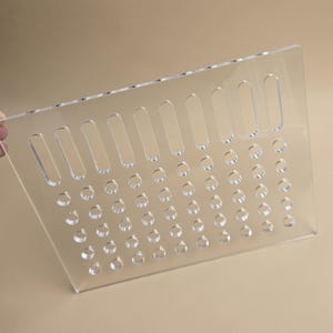 May include: A transparent acrylic sheet featuring a pattern of small, circular holes and larger, oblong cutouts. The sheet is held at an angle, highlighting its clarity and the precise cut patterns. The background is a neutral beige colour.