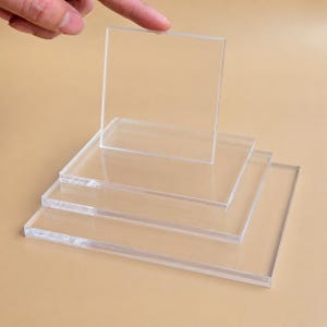 May include: Clear acrylic square sheets, stacked and arranged. One sheet is lifted by a finger. The sheets are transparent and have a smooth surface. The image is taken against a neutral background.