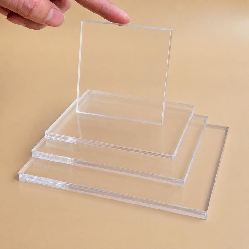 May include: Clear acrylic square sheets, stacked and arranged. One sheet is lifted by a finger. The sheets are transparent and have a smooth surface. The image is taken against a neutral background.