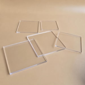 May include: Several clear, rectangular acrylic sheets of varying sizes are arranged on a neutral-toned surface. The transparent material allows the background color to show through, creating a subtle interplay of light and shadow. The sheets are slightly overlapping.