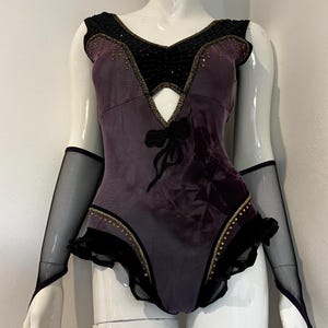 May include: A plum-colored bodysuit with black accents and a keyhole design. The bodice features gold beading and a black bow. Includes matching black mesh arm sleeves. The garment is displayed on a mannequin.