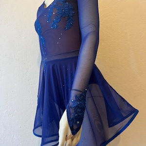 Könnte beinhalten: Königsblaues Eiskunstlaufkleid mit langen, transparenten Ärmeln und einem ausgestellten Rock. Das Oberteil hat aufwendige Blumenstickereien und funkelnde Verzierungen. Das Kleid ist für Auftritte und Wettbewerbe konzipiert.