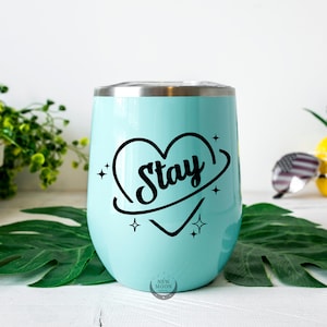 以下が含まれることがあります： 「Stay」と書かれた黒いハートのデザインが施された、薄い青色のステンレス製タンブラー。ハートは黒い円に囲まれ、両側に星が描かれている。
