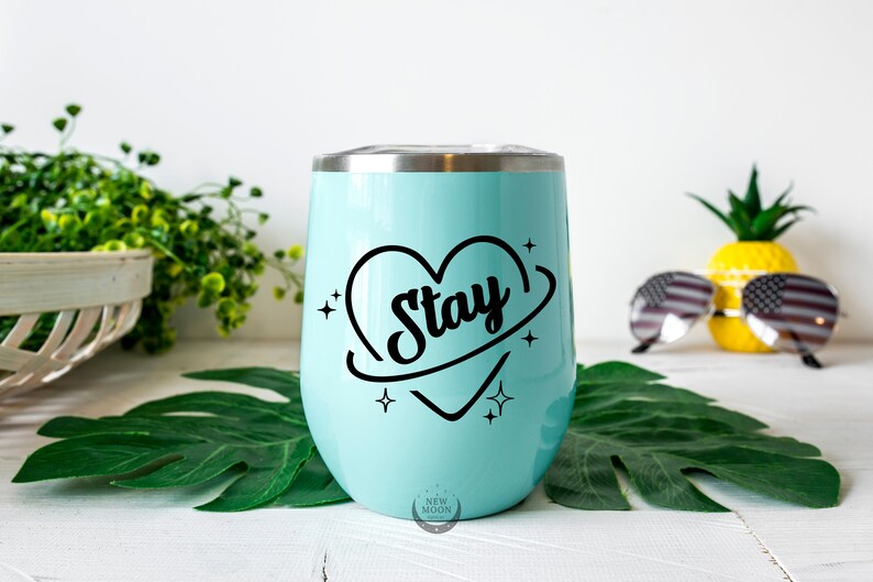 以下が含まれることがあります： 「Stay」と書かれた黒いハートのデザインが施された、薄い青色のステンレス製タンブラー。ハートは黒い円に囲まれ、両側に星が描かれている。