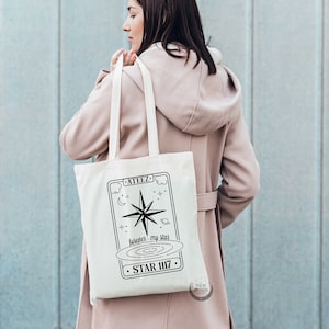 以下が含まれることがあります： 星、月、そして「GALAXY」、「between my stars」、「STAR III7」というテキストが描かれた白地のキャンバス地のトートバッグです。黒と白のグラフィックデザインが施されています。