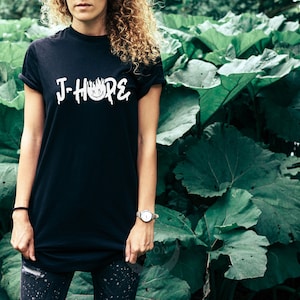 Może przedstawiać: Czarny T-shirt z białym napisem "J-Hope" i stylizowanym wzorem płomienia. Osoba nosząca koszulkę stoi przed zielonym, liściastym tłem.