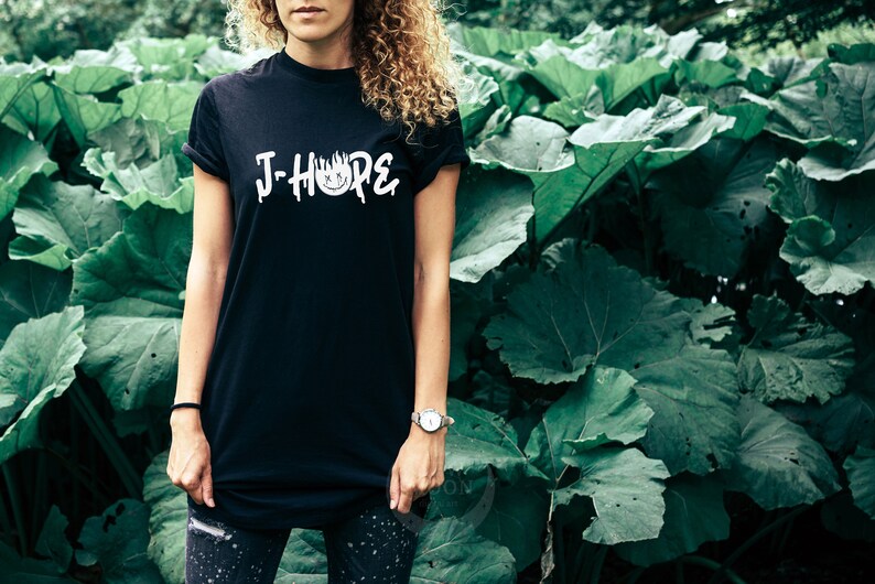 Może przedstawiać: Czarny T-shirt z białym napisem "J-Hope" i stylizowanym wzorem płomienia. Osoba nosząca koszulkę stoi przed zielonym, liściastym tłem.