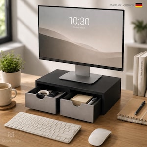 Op de afbeelding: Een zwarte monitorstandaard met twee lades met lichtgrijze fronten. De standaard ondersteunt een computermonitor die de tijd 10:30 en de datum maandag 20 mei weergeeft. De tekst Made in Germany is zichtbaar in de rechterbovenhoek.
