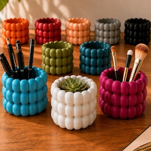 Op de afbeelding: Een collectie cilindrische houders met een bubbeltextuur aan de buitenkant, verkrijgbaar in diverse kleuren waaronder oranje, rood, perzik, grijs, zwart, olijfgroen, teal, blauw, wit en magenta, voor het opbergen van pennen en penselen.