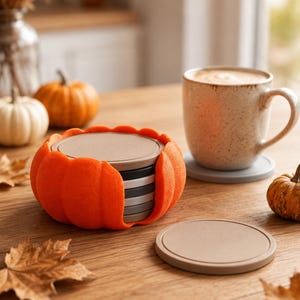 Puede incluir: Un juego de posavasos circulares apilados dentro de un soporte naranja con forma de calabaza. Los posavasos son de varios tonos de gris, marrón y beige. Un posavasos yace plano sobre la superficie junto a una taza de cerámica, mientras otros siguen en el soporte.