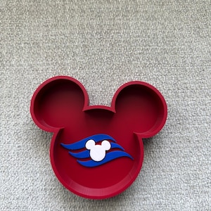 May include: A red Mickey Mouse-shaped tray with two circular ears and a circular base. The tray features a white Mickey Mouse head and blue waves design. The tray is likely made of plastic or a similar material.
